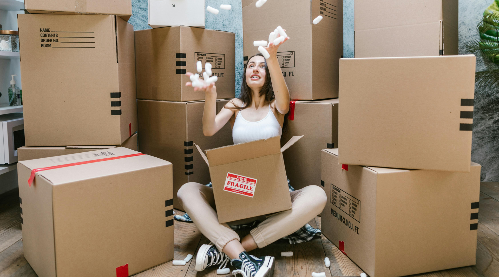 Woman sits among moving boxes, happily tossing items into the air. Boxes are stacked in a room with a bright background.