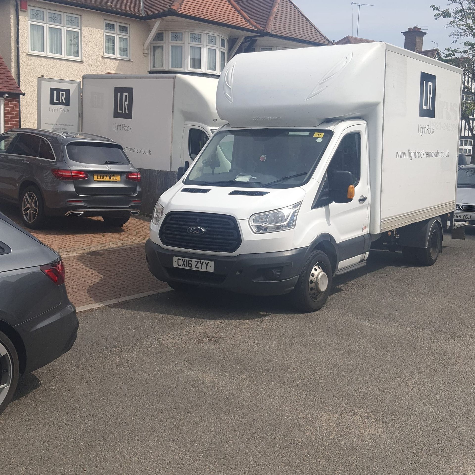 White moving van and another truck parked on a driveway with cars next to it. The van has