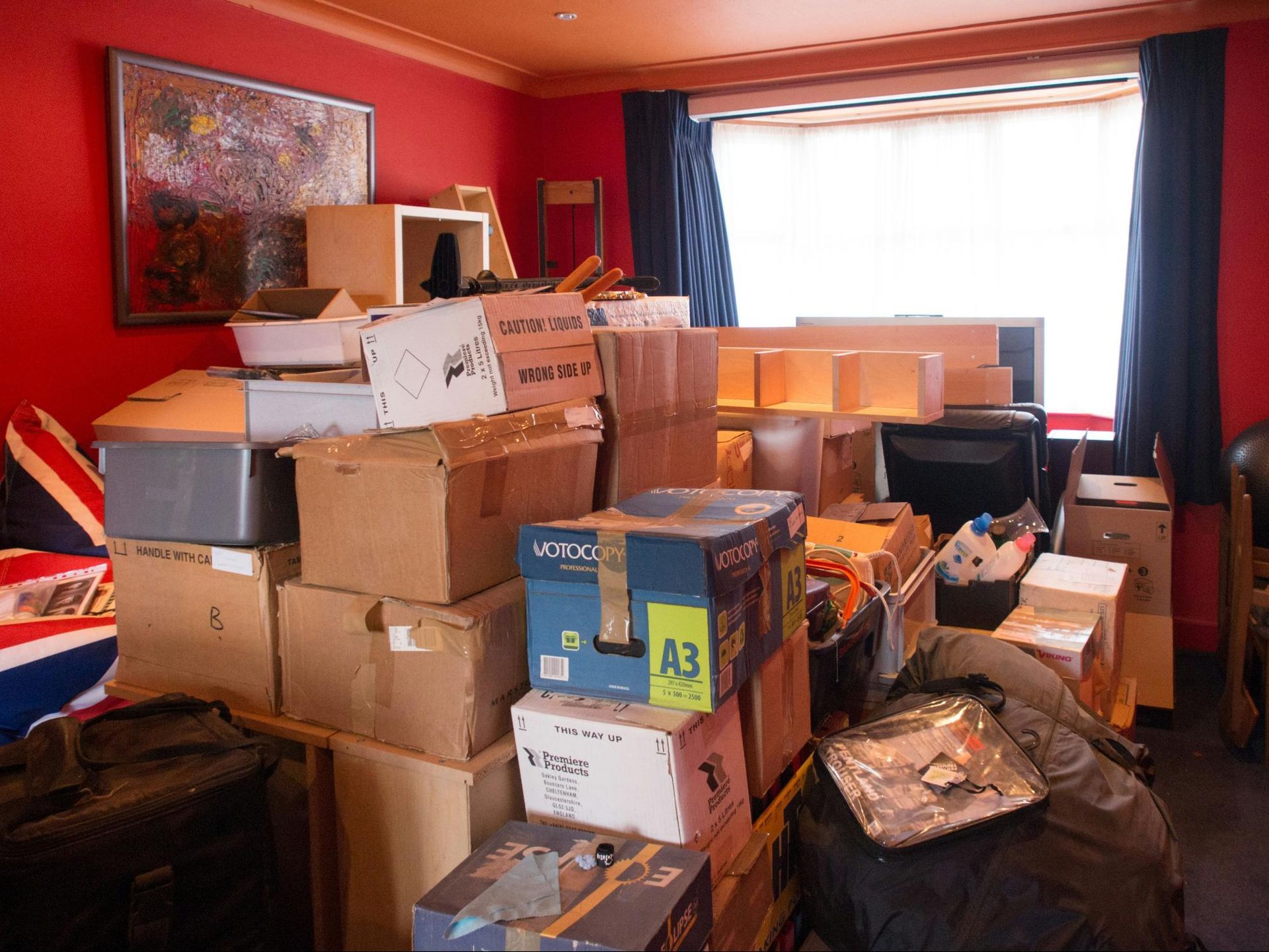 Boxes fill a red-walled room near a window. Moving supplies create a cluttered scene.