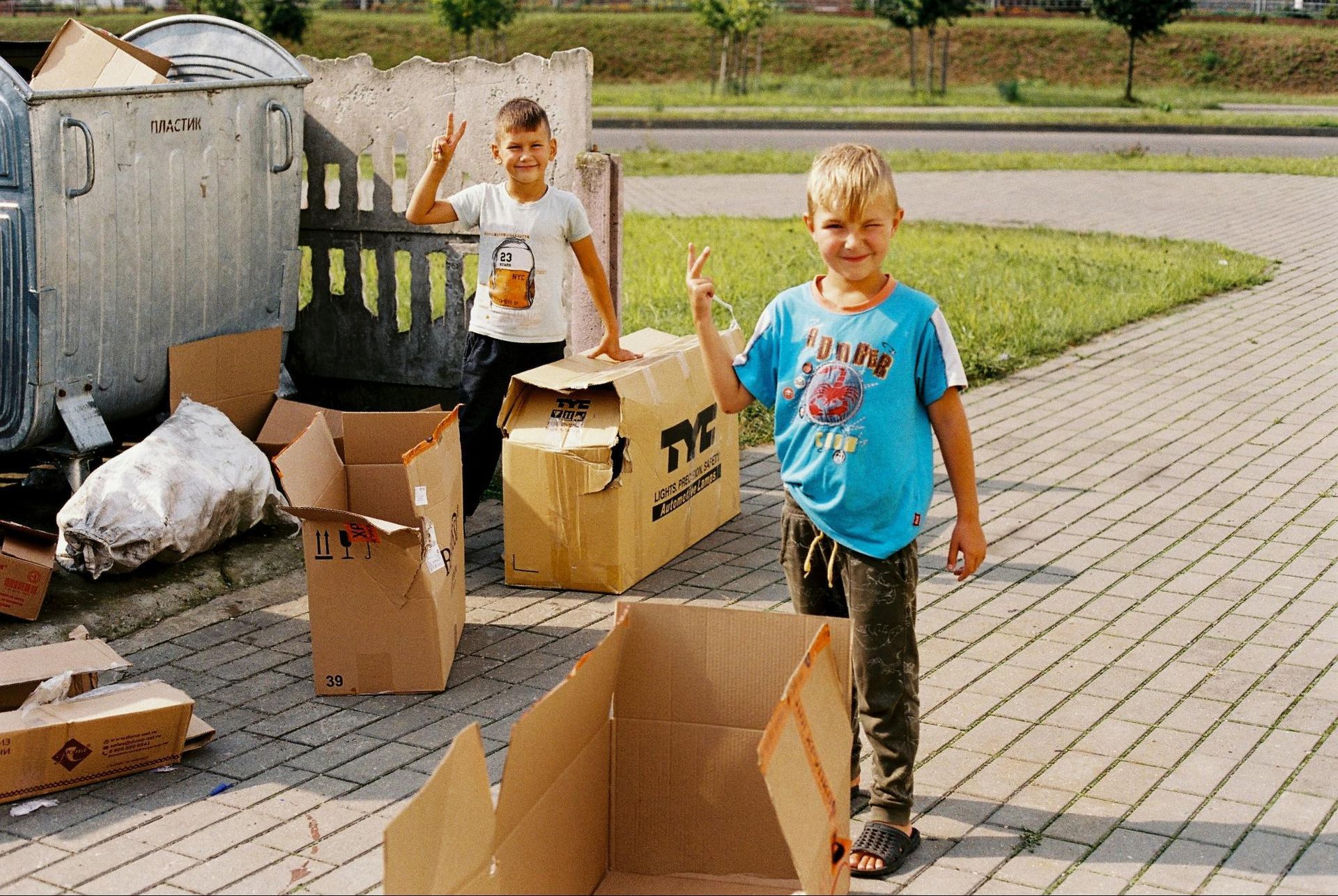 Two children posing by cardboard boxes and trash bins, giving peace signs.