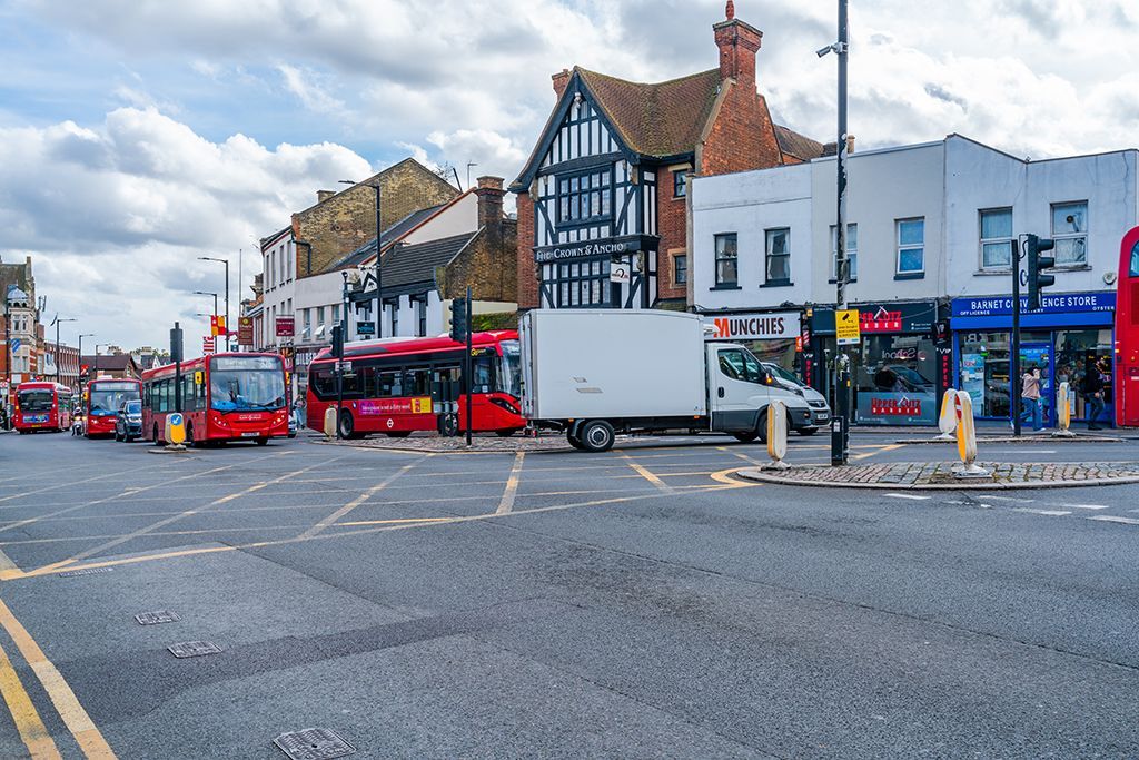 Busy street scene in London, England. Red buses and a white van are on the road. Buildings with shops line the street.