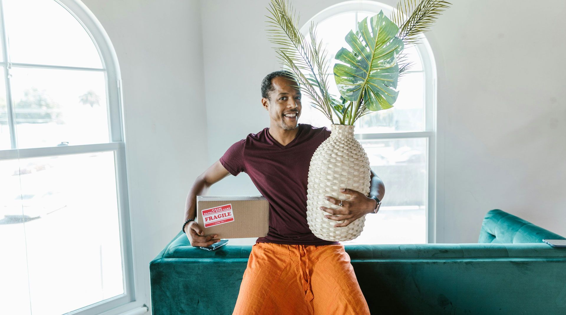 Man in orange pants and maroon shirt holds a large vase with plants and a box labeled 
