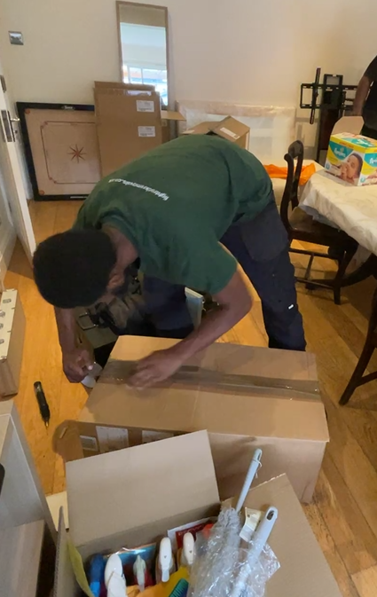 A person wearing a green shirt and black pants, kneeling, cutting open a cardboard box on a wood floor.
