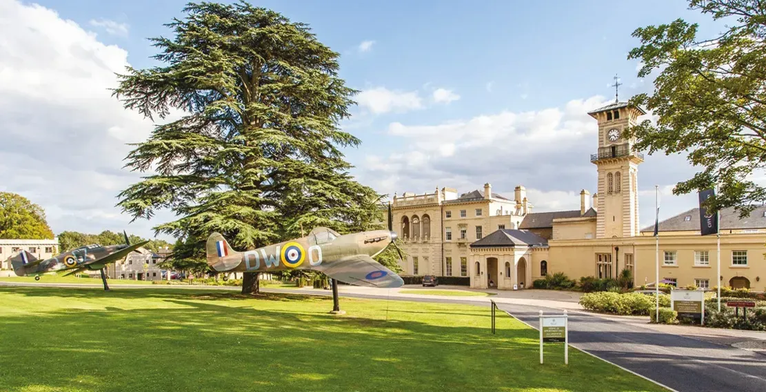 A historic yellow building with a clock tower, airplane on lawn, green trees, sunny day.