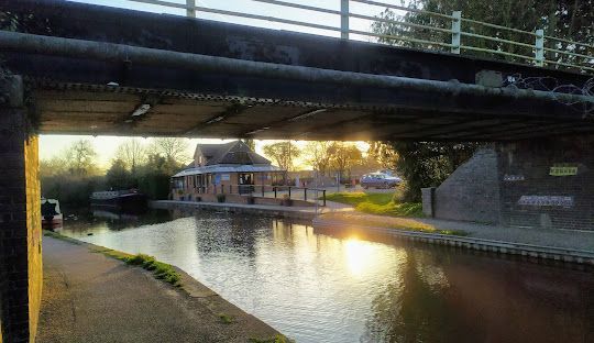 A canal with a floating building under a bridge at sunset. Golden light reflects on the water.