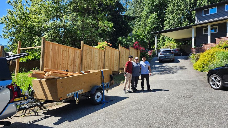 Workers beside a trailer loaded with wooden fence panels on a driveway near a house