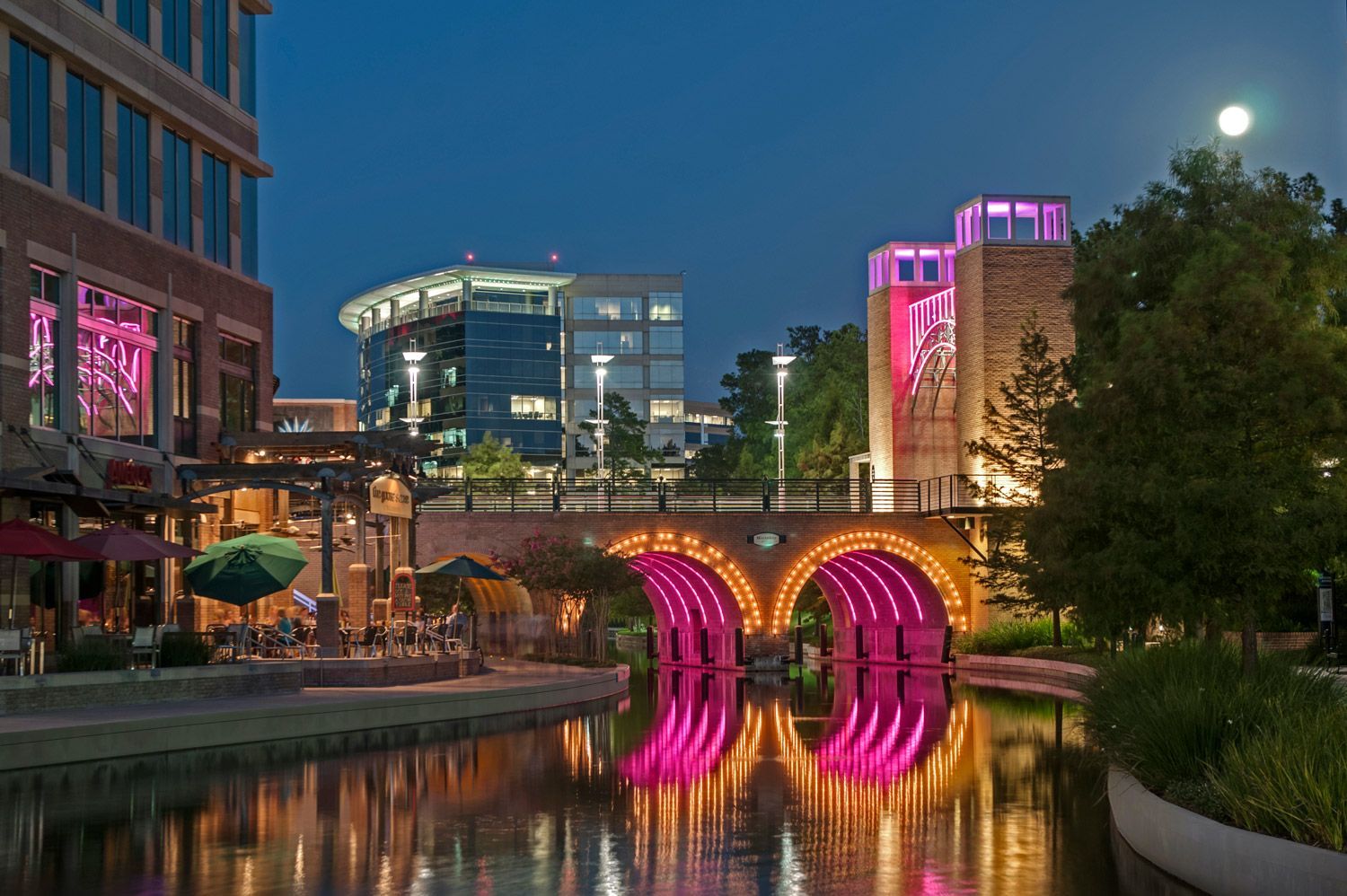 A bridge in The Woodlands over a river with buildings in the background at night