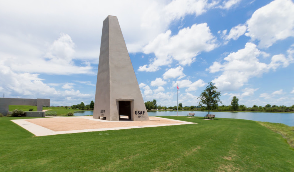 A large obelisk in Sugar Land is sitting in the middle of a grassy field next to a lake.
