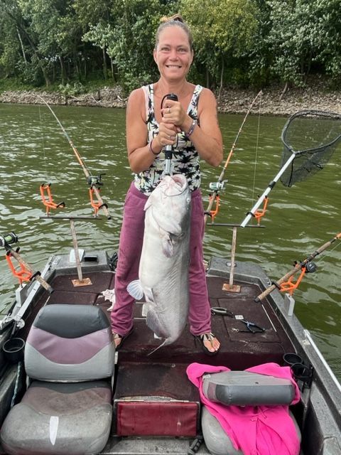 Woman on a boat holding a large catfish, showing it off. Fishing rods and a net are visible.