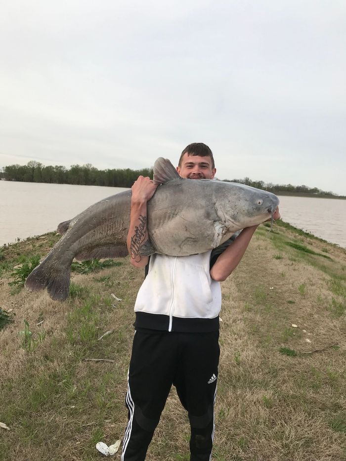 Man holding a very large catfish. He is smiling near a body of water on a cloudy day.