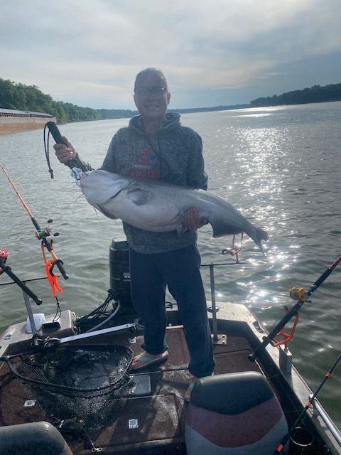 Man on a boat holding a large blue catfish, with rods, net, and river in the background.