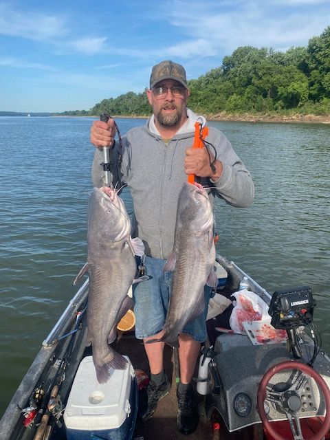 Man on boat holding two large catfish he caught. Blue water, trees in background.