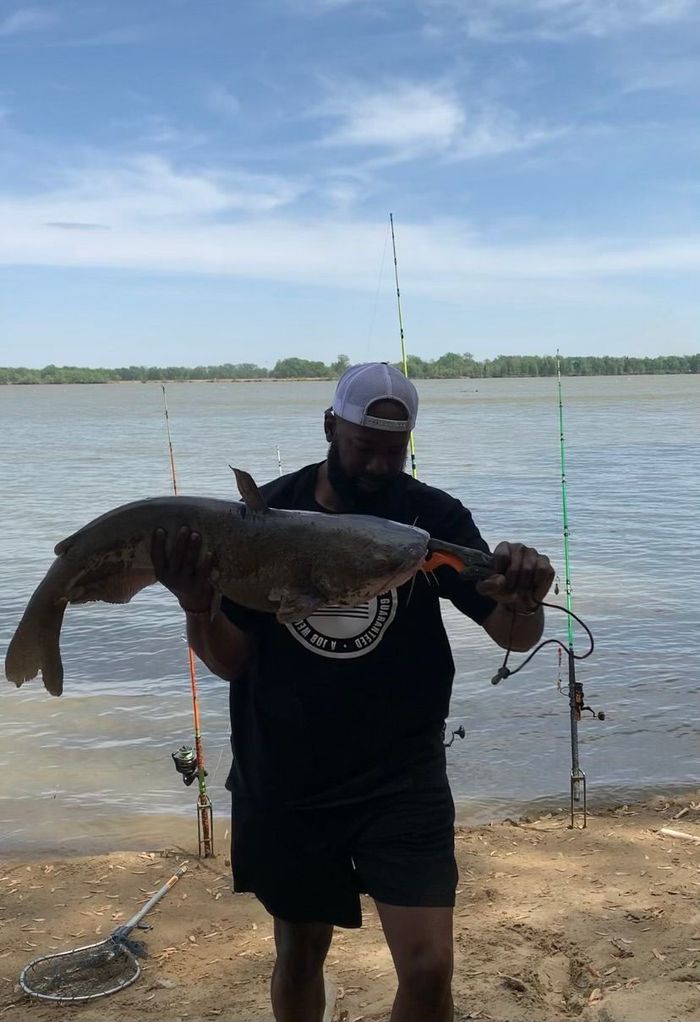Man holding a large catfish near a body of water, fishing poles visible. Cloudy sky.