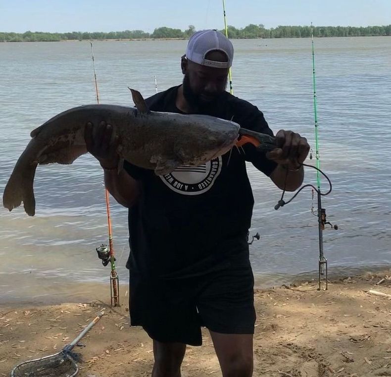 Man holding a large catfish by the water, fishing poles in the background.
