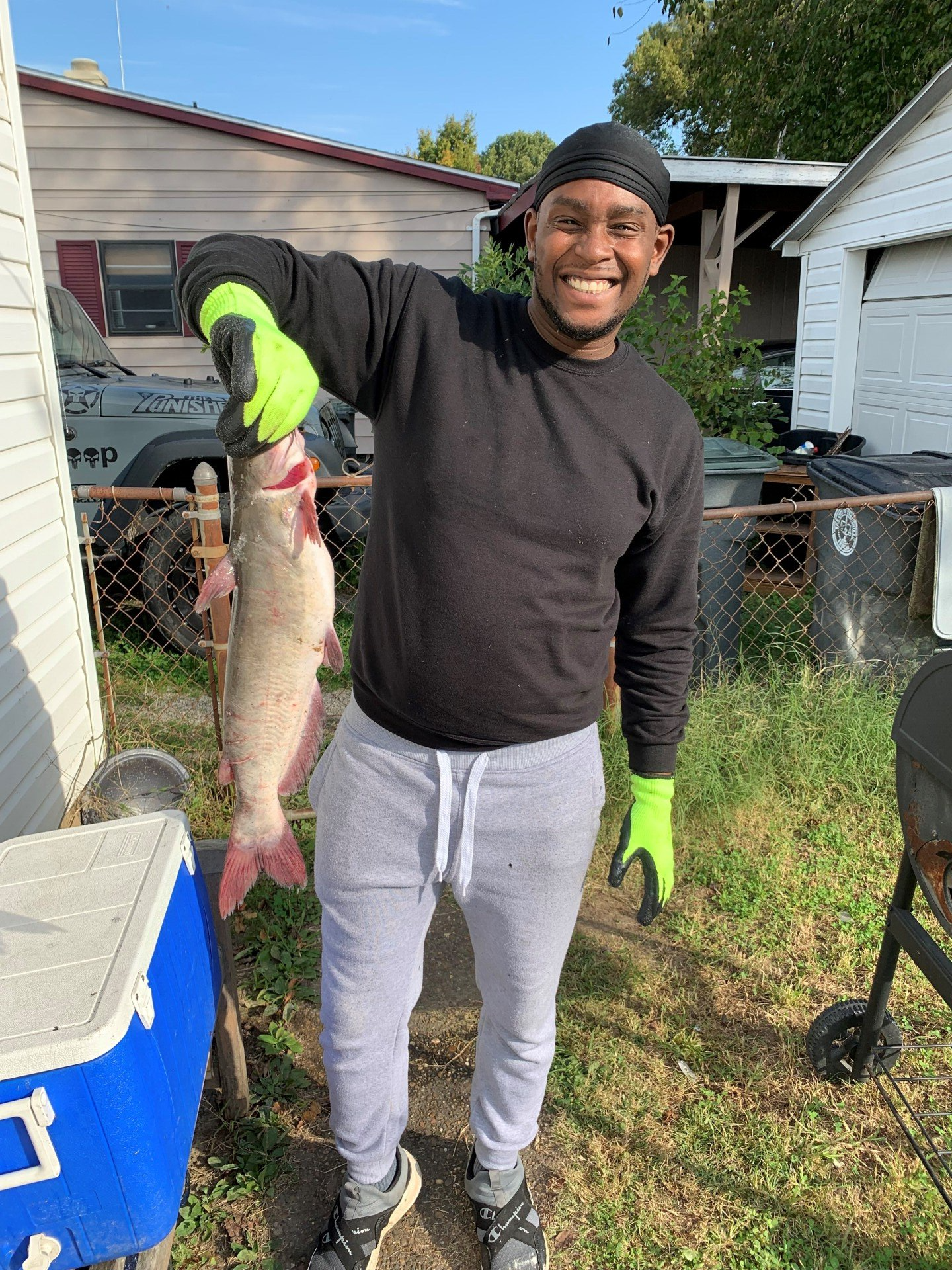 Man holding a fish outside, wearing black sweatshirt, gray sweatpants, green gloves. Smiling.
