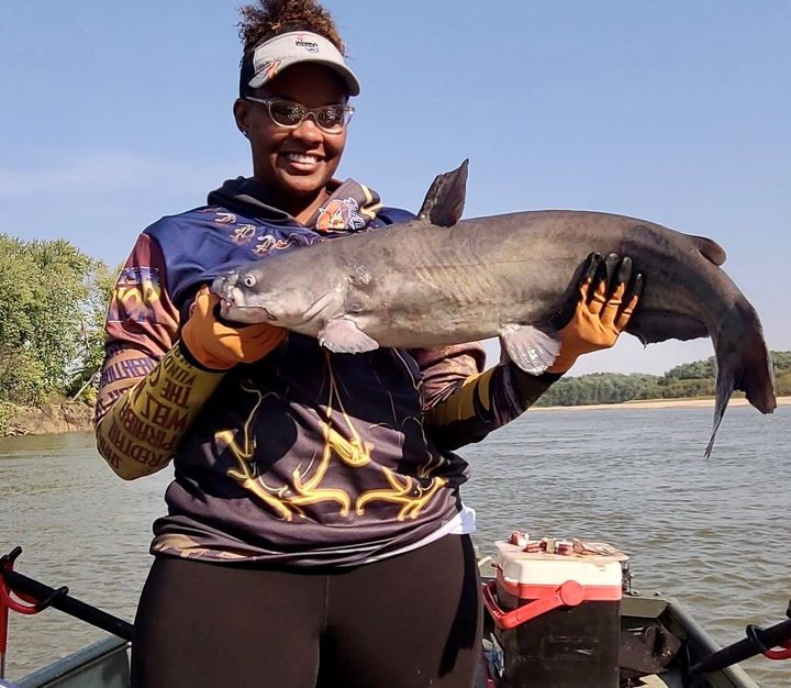 Woman holding a large catfish on a boat, smiling, wearing glasses and fishing apparel. River background.