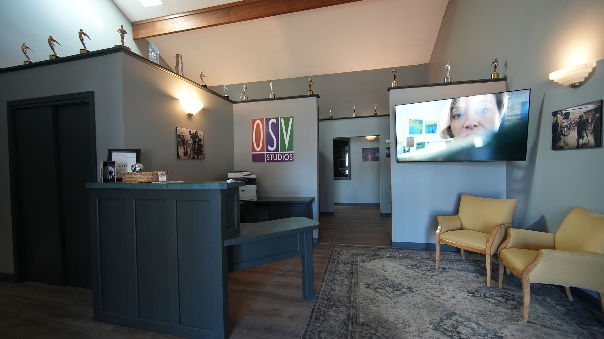 Modern office lobby with reception desk, gray walls, TV screen, and two yellow chairs on a rug