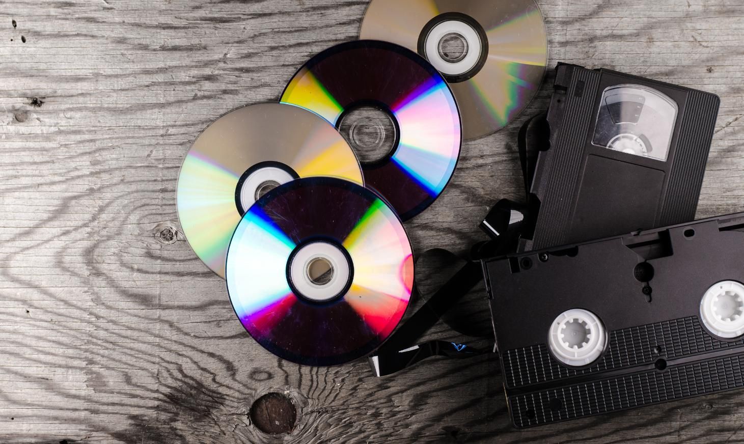 A wooden table topped with a VHS tape, CDs and DVDs.