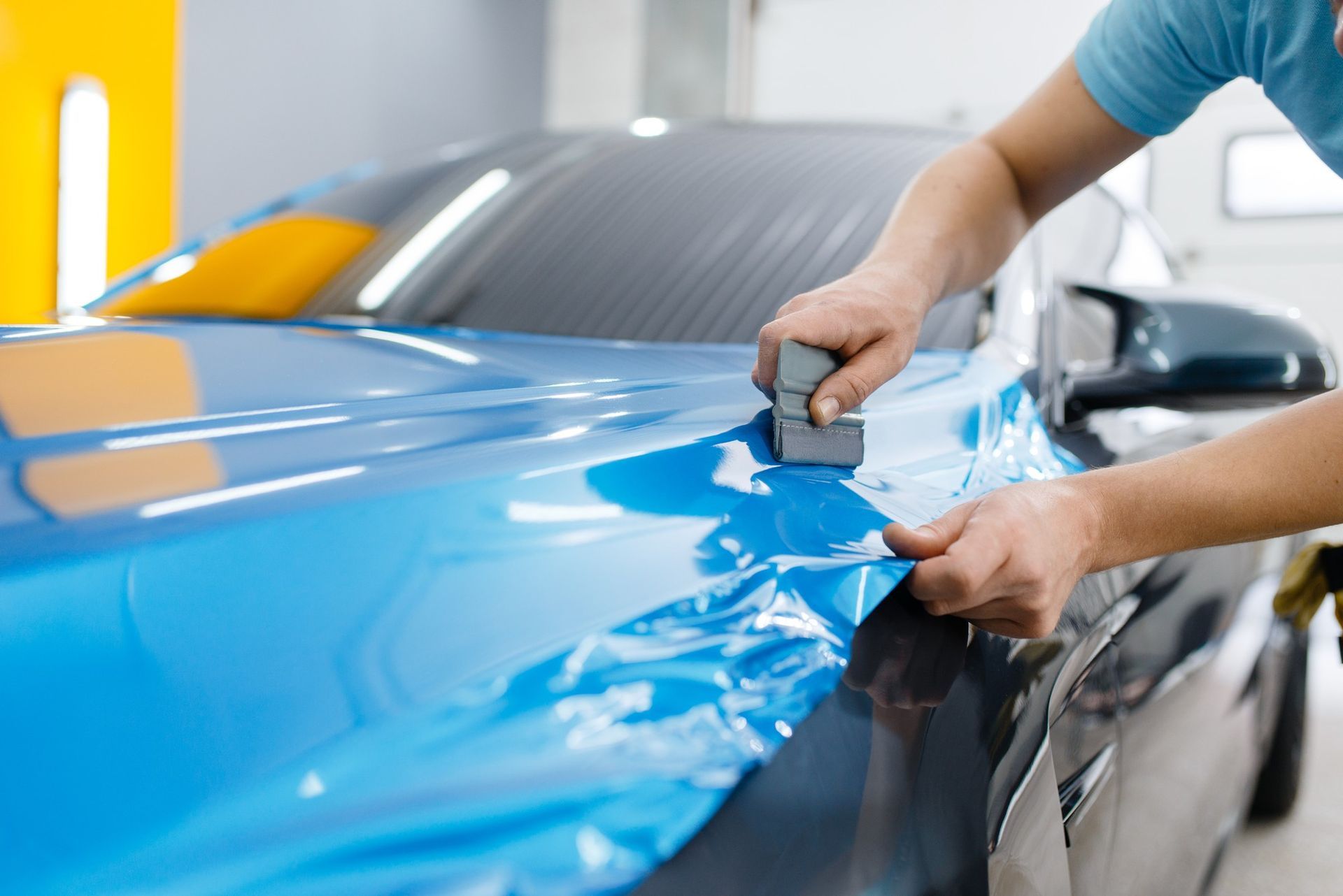 A worker uses a squeegee to apply blue vinyl wrap to a car hood in a workshop.