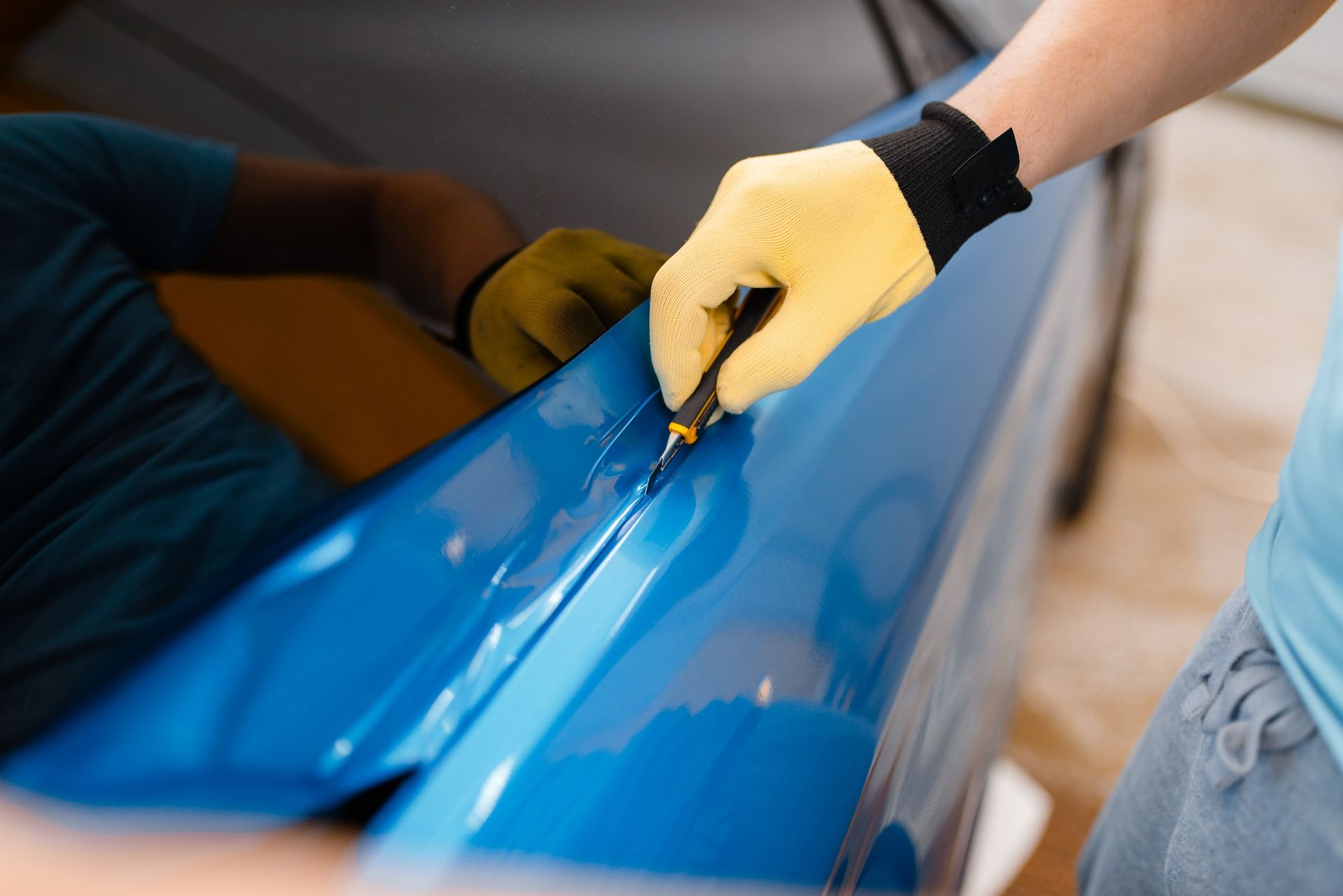 A gloved hand uses a precision knife to trim excess material along the edge of a blue car door.