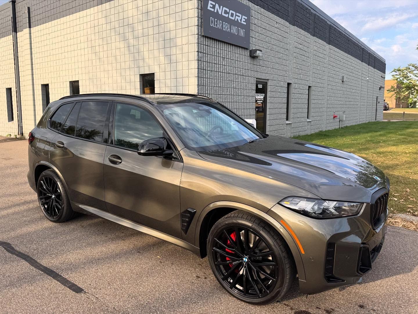 A dark bronze BMW X5 SUV parked outside a building with a light-colored brick exterior.