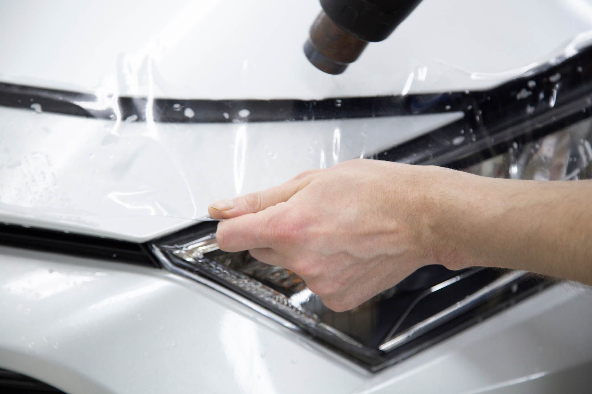 A hand uses a heat gun to apply a protective clear film onto the white hood of a car.