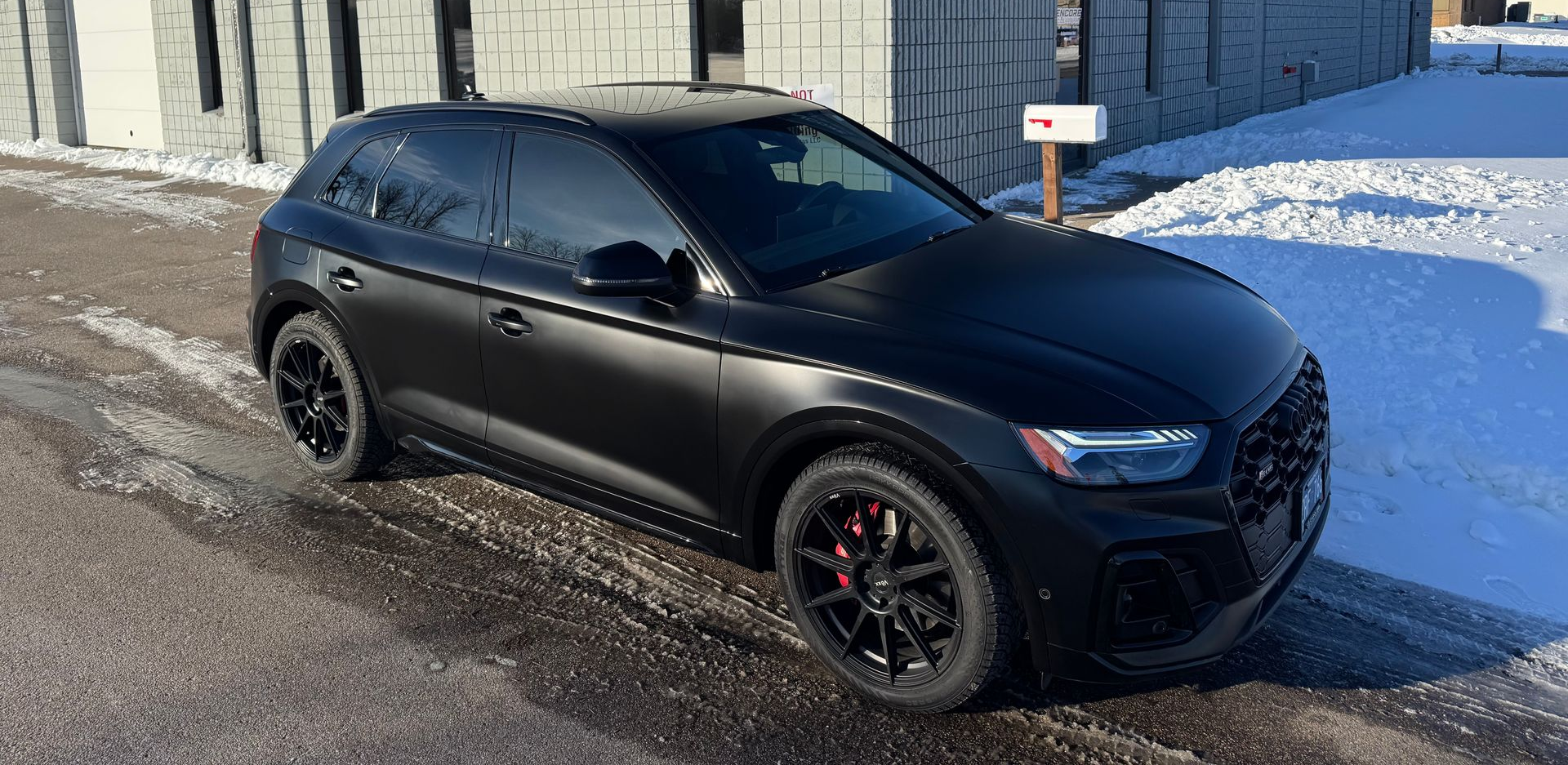 A matte black Audi SUV with black rims and red brake calipers parked on a gravel shoulder next to snow.