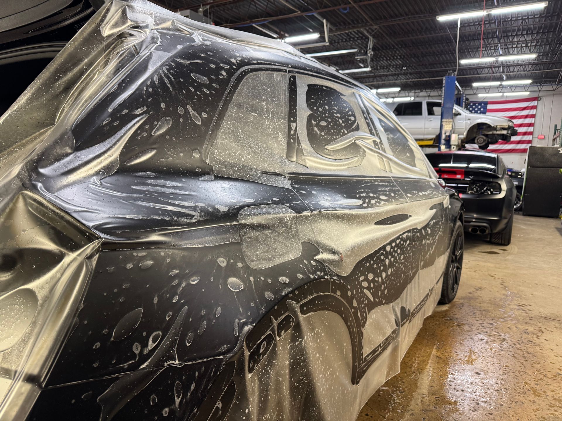 A black car covered in soapy water being detailed inside a garage, with another vehicle and an American flag visible.
