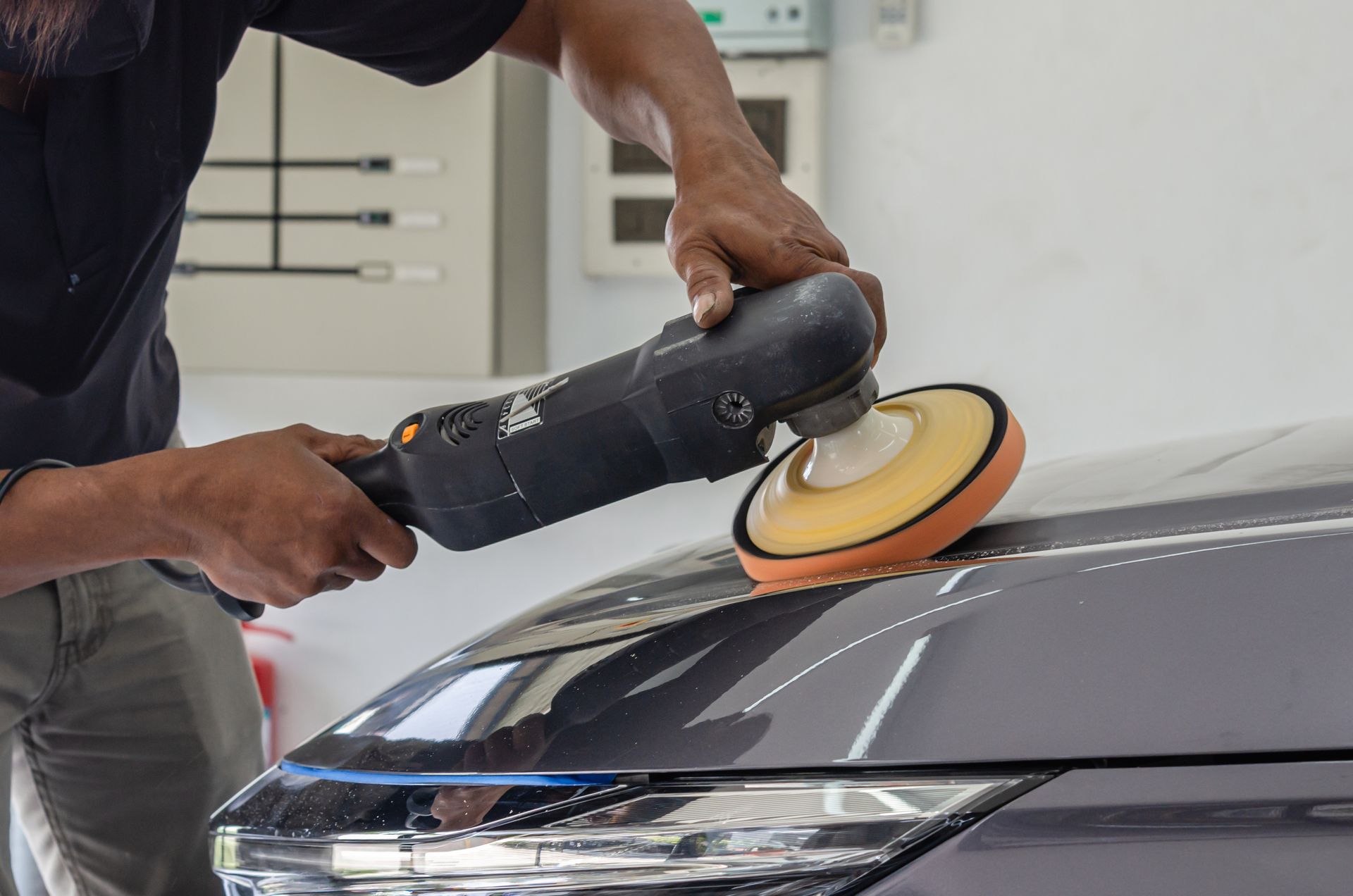 A person uses a rotary buffer with an orange foam pad to polish the hood of a gray car in a workshop.