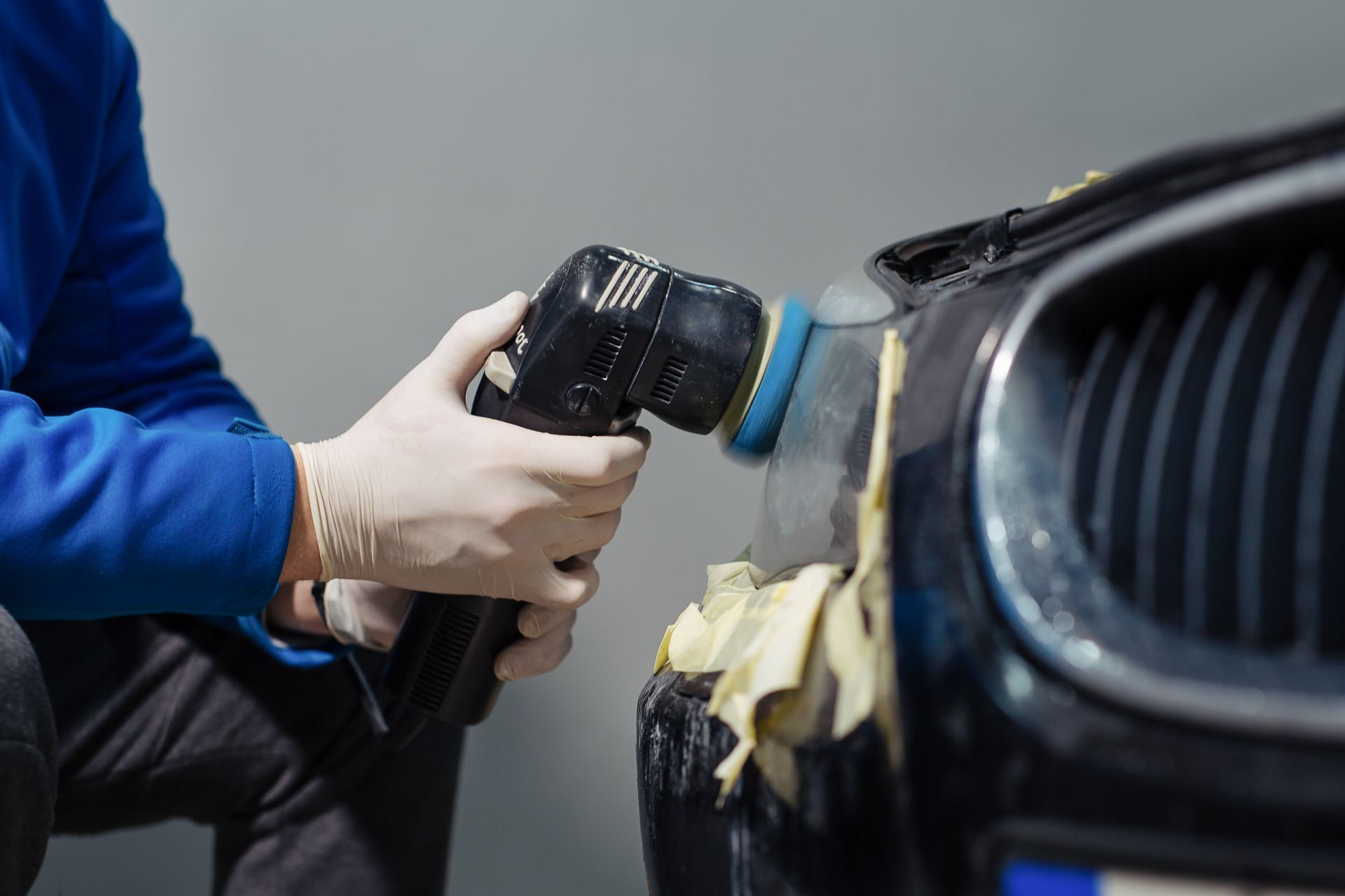A person wearing white gloves uses a power polisher on the front of a dark car with masked-off trim.