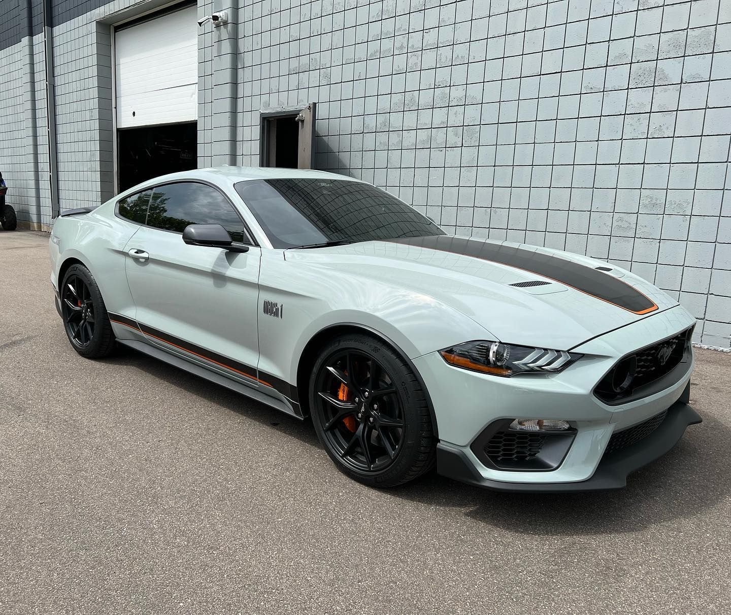 A light gray Ford Mustang Mach 1 with black racing stripes and black wheels parked in front of a gray brick building.