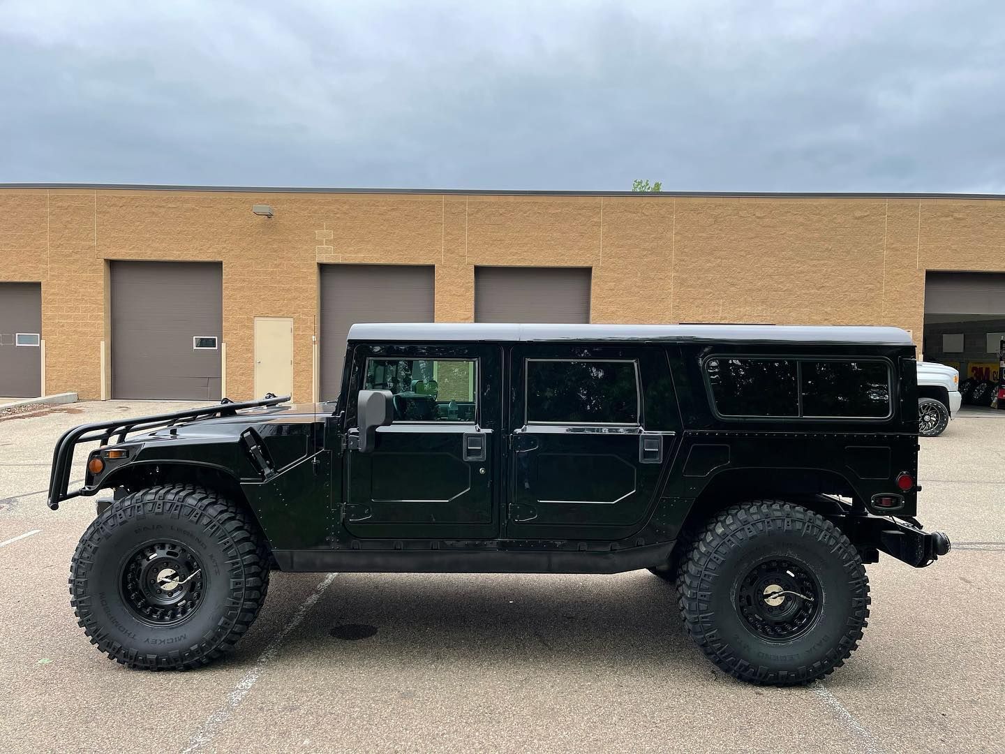 A side view of a black Hummer H1 parked on asphalt in front of a tan industrial building.