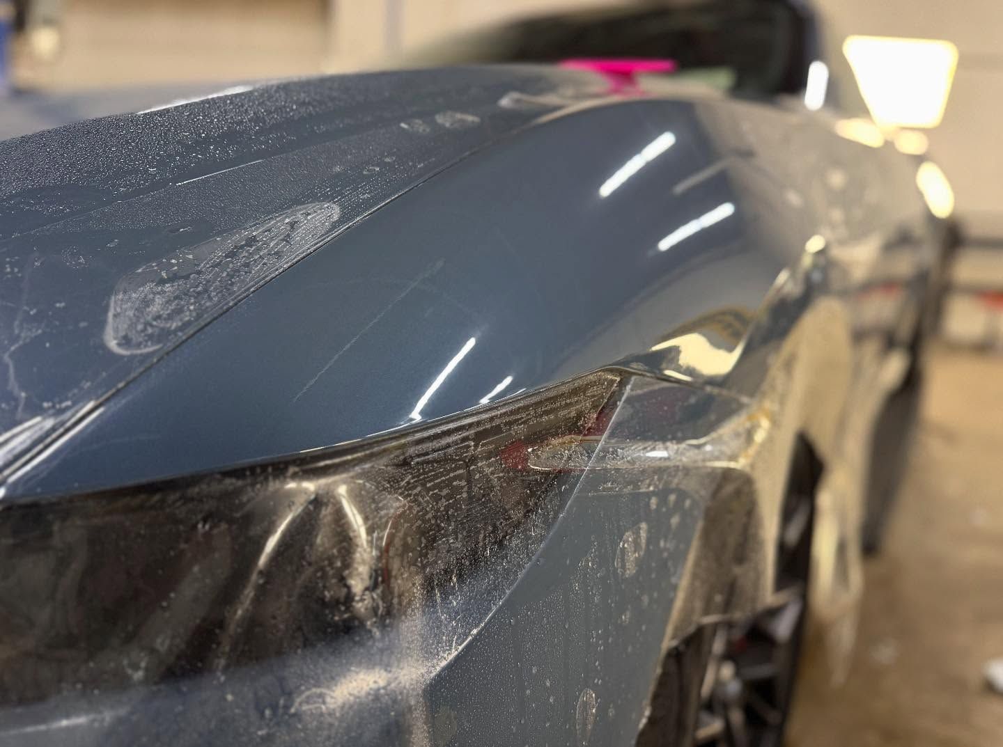 A close-up of a blue car hood and headlight during a paint protection film installation in a shop.