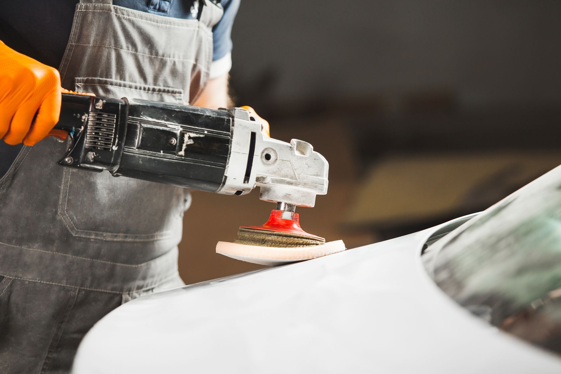 A person in grey work overalls and orange gloves uses a power polisher on a white car hood.