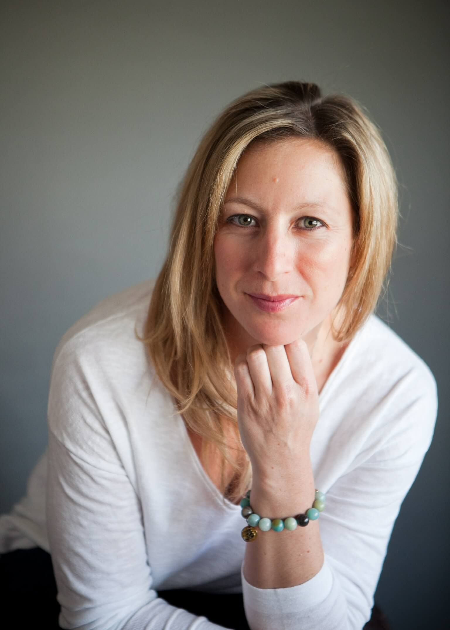 Woman in white top, resting her chin on her hand against a gray background.