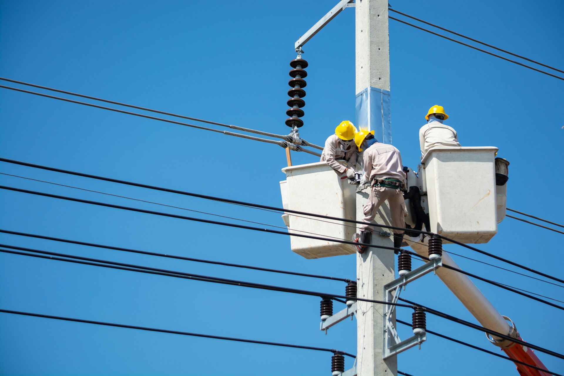 Utility workers in bucket lifts repairing power lines on a utility pole under a blue sky