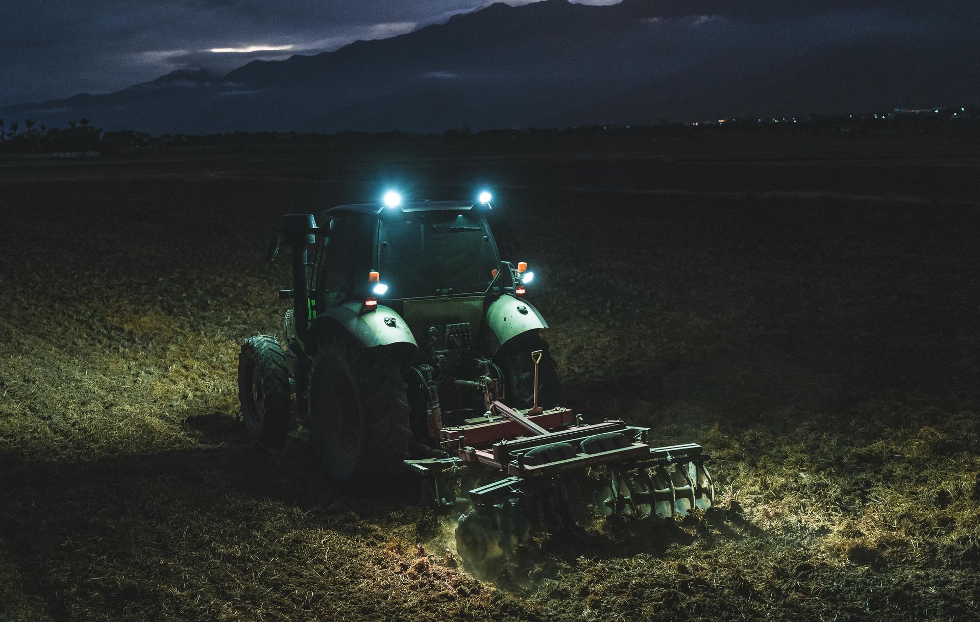 Tractor with bright headlights working in a dark field at night, attached to farming equipment