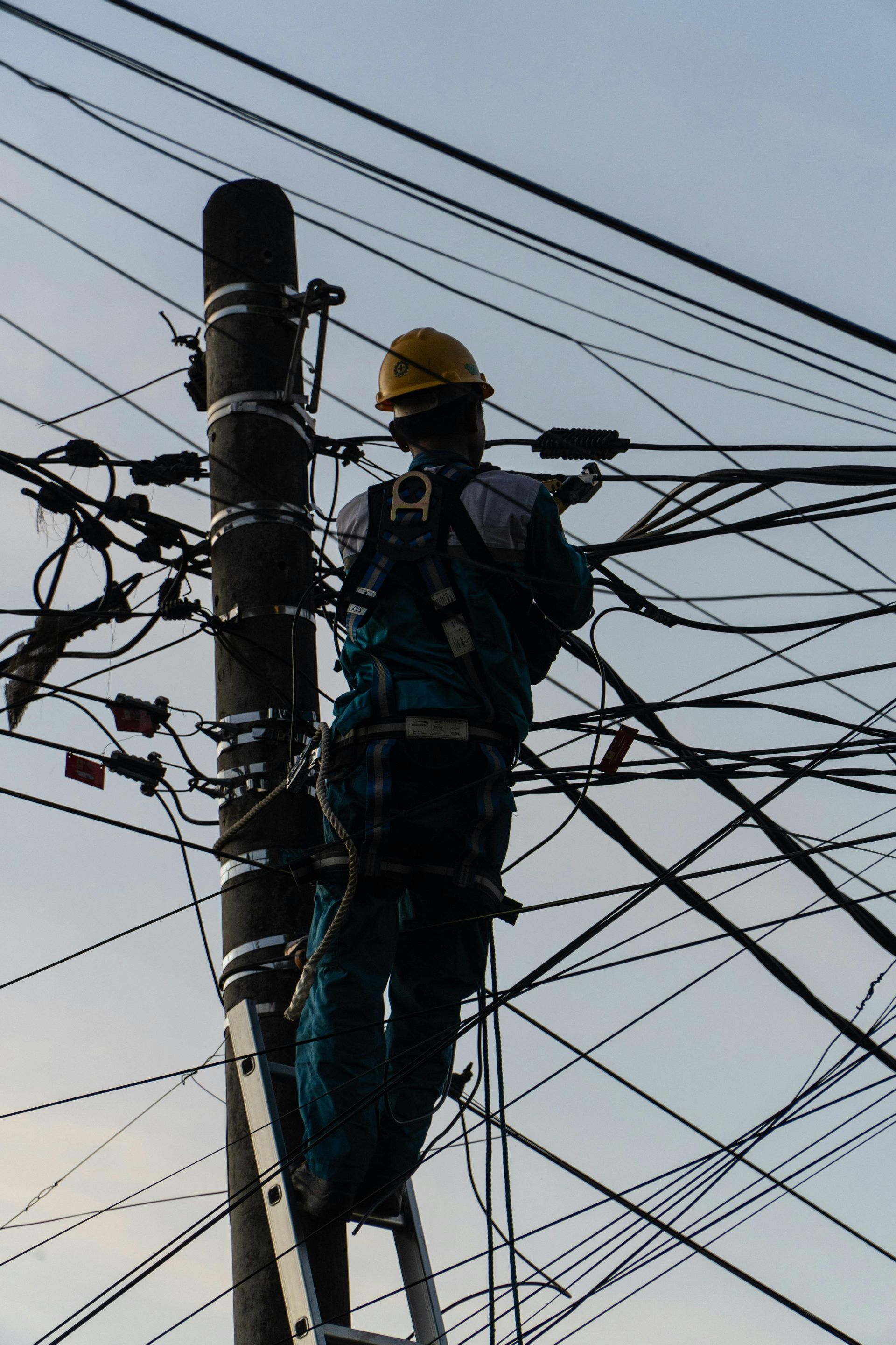 Utility worker in yellow hard hat climbing a power pole amid tangled overhead wires