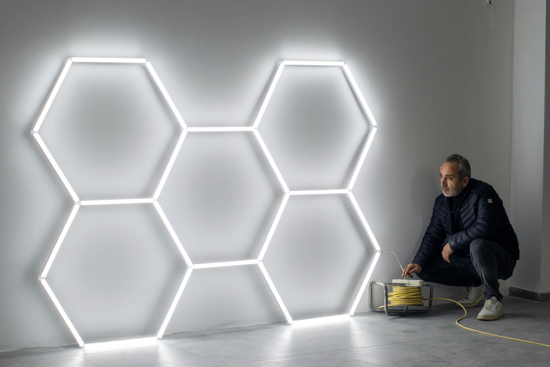 Man kneels beside glowing hexagonal wall lights in a gray room, adjusting a power source.