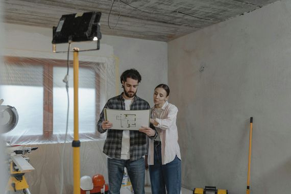 Two people reviewing a floor plan in an unfinished room with construction lights and tools.