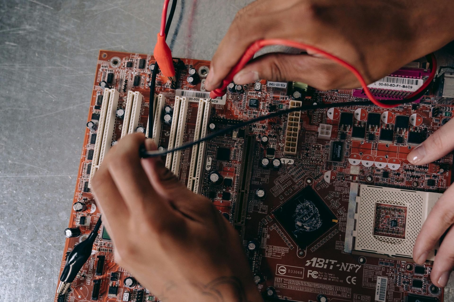 Hands probing a circuit board with red test leads and tweezers on a workbench