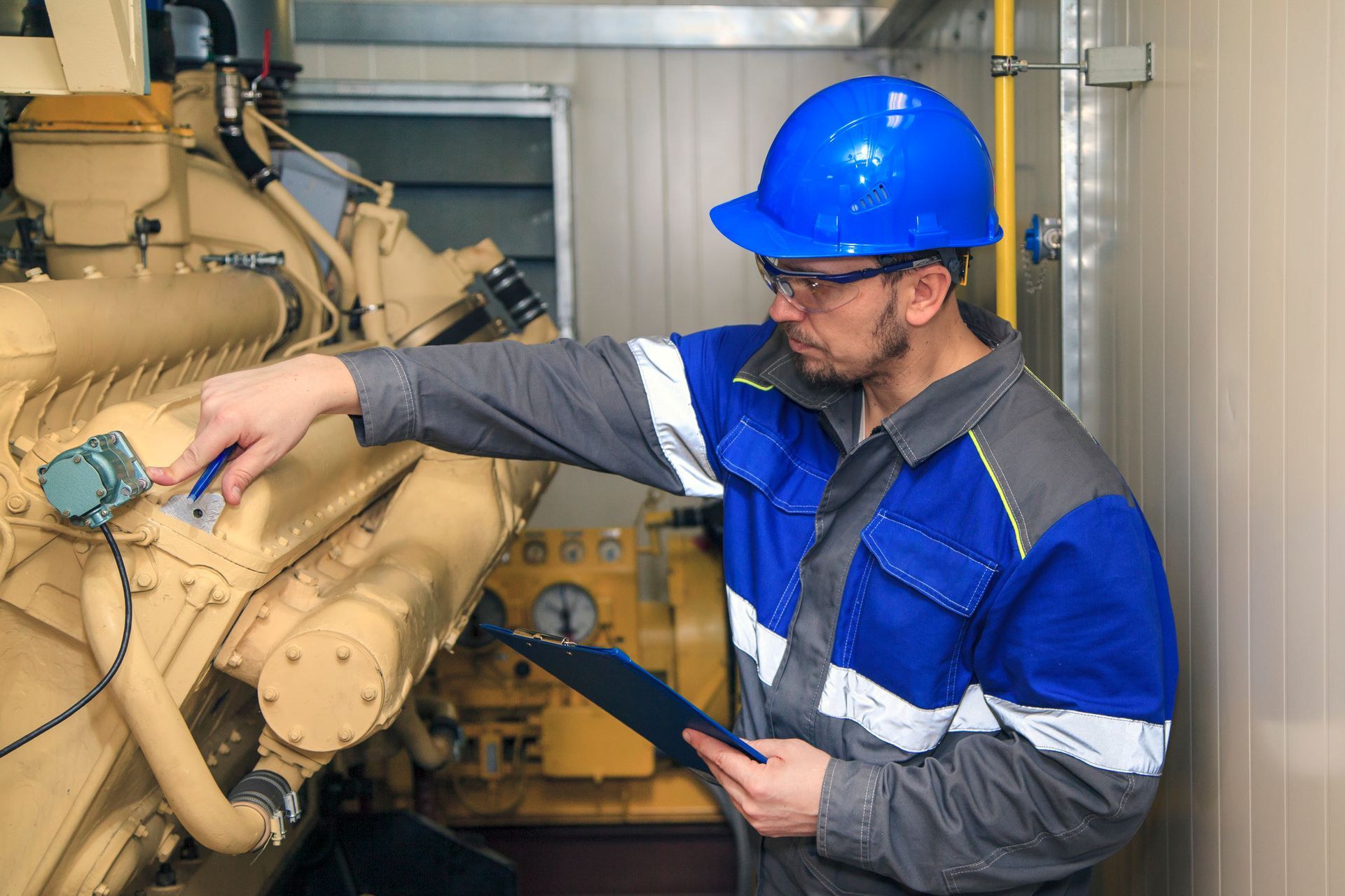 Technician in blue hard hat inspecting a large industrial engine with a tablet in a machinery room