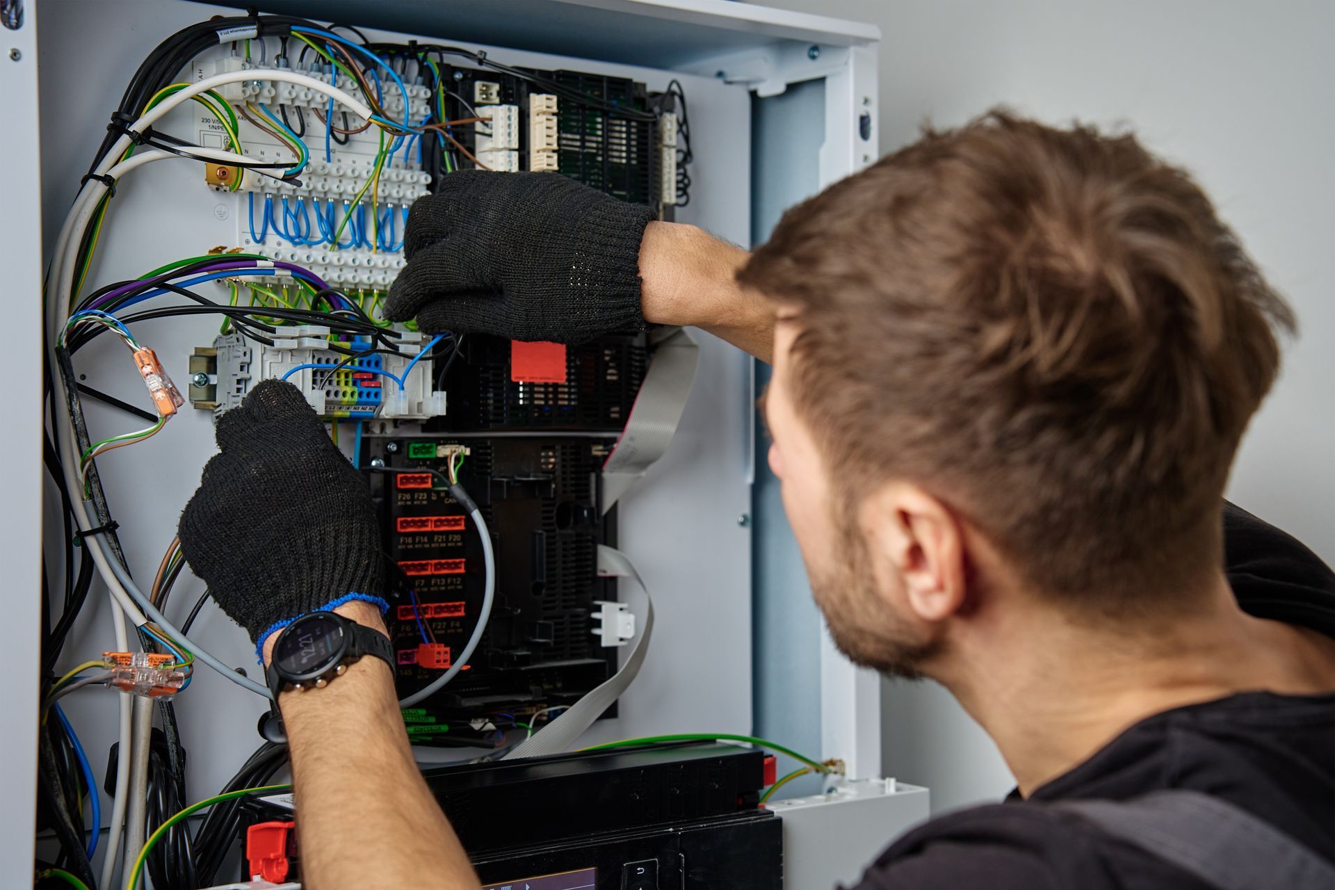 Technician wearing gloves working inside an open electrical control panel with tangled wires.