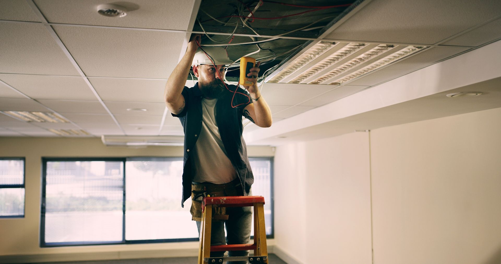 Worker standing on a ladder fixing a dropped ceiling tile in an office room