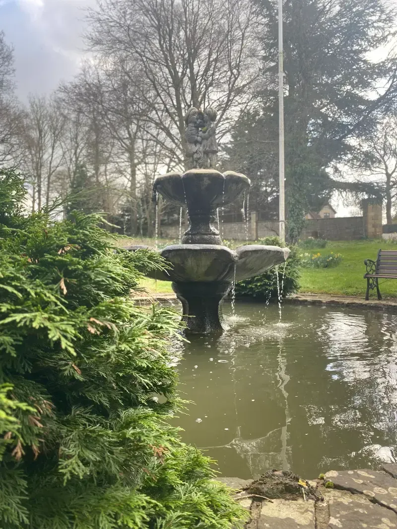 Woodlands Manor Care Home | A fountain is in the middle of a pond in a park.