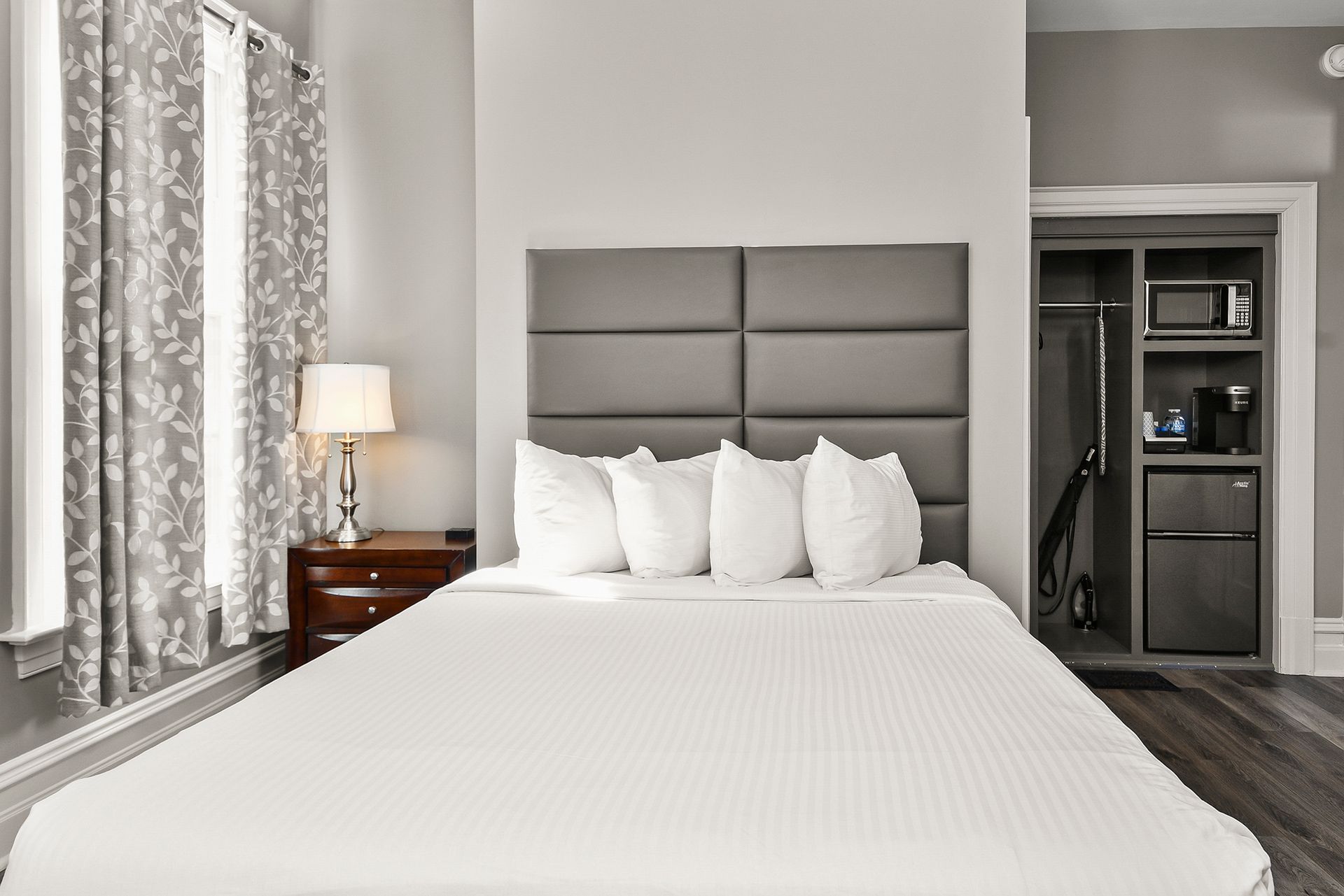A tidy bedroom with a white-covered bed, grey tufted headboard, bedside lamp, and a small kitchenette in a wall nook.