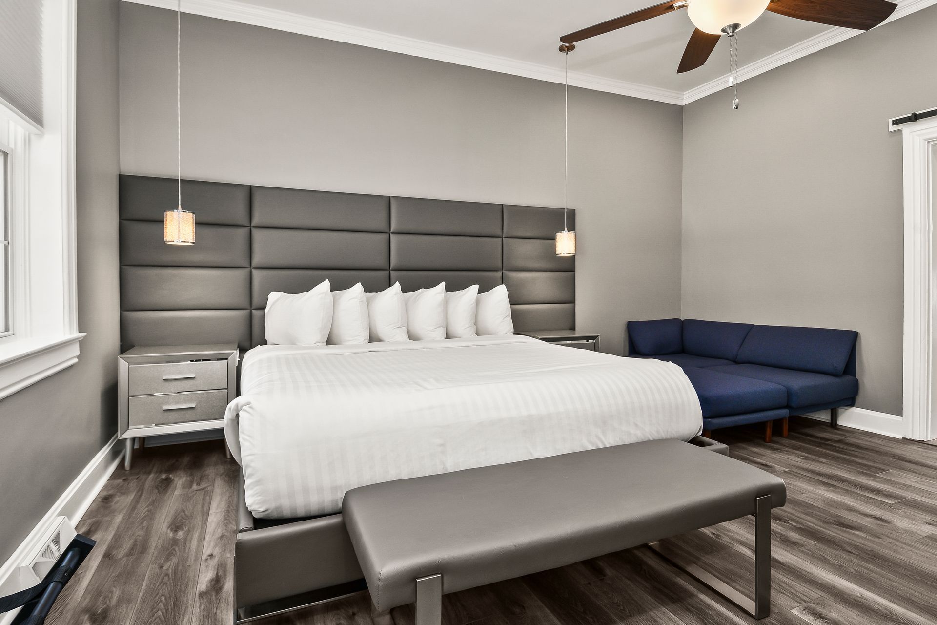 A hotel room with a large bed against a tall, gray upholstered headboard, a navy blue sofa, and a gray bench at the foot.