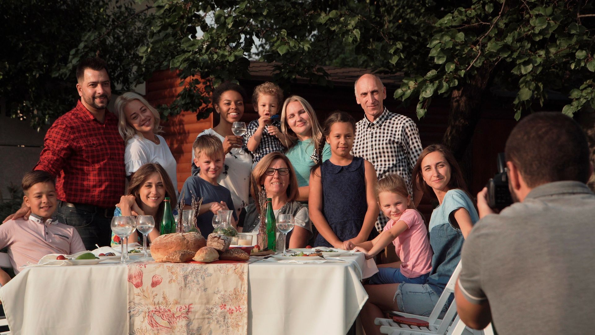 A group of people gathers outdoors around a table covered in a white cloth while one person photographs them.