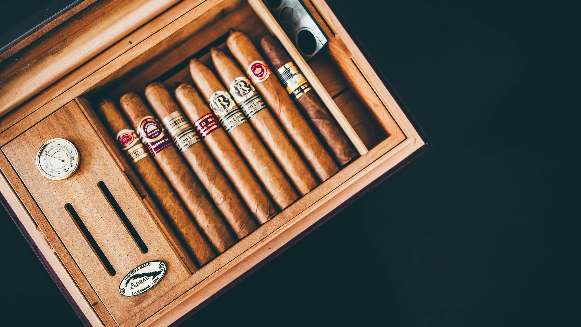 A wooden humidor containing eight premium cigars arranged in a row, with an analog hygrometer visible on the side.