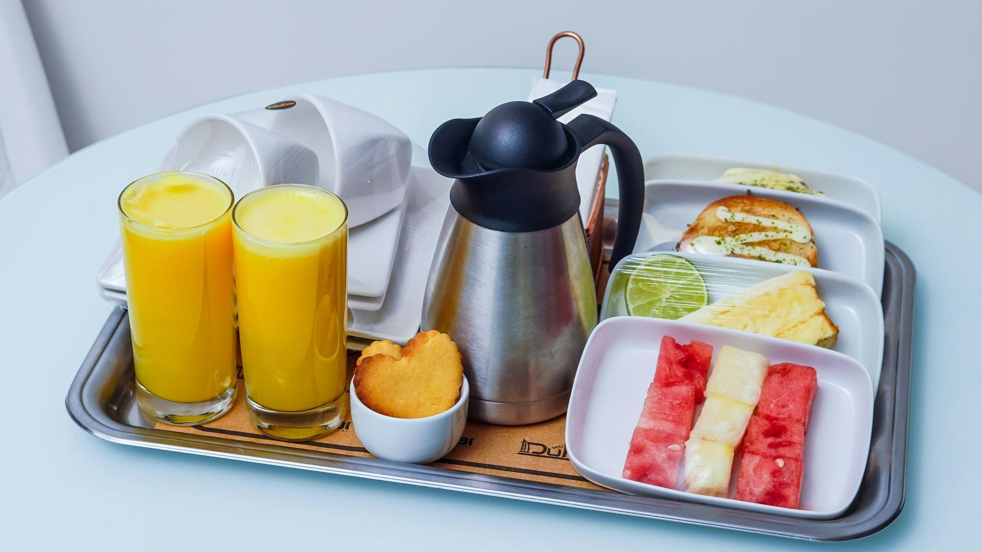 A silver tray holds two glasses of orange juice, a coffee carafe, a bowl with a pastry, and plates of fruit and toast.